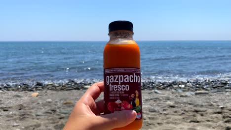 Holding-a-fresh-traditional-gazpacho-mini-bottle-from-Spanish-Mercadona-supermarket-at-the-sunny-beach-in-Estepona-Spain-with-sea-view,-cold-soup-on-the-go,-4K-static-shot