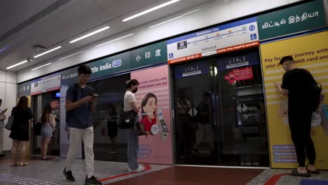 Pendler-Und-Singapurische-Einheimische-Warten-Neben-Den-Bahnsteigtüren,-Während-Sich-Der-MRT-Zug-Am-Bahnhof-Little-India-DT12-Nähert