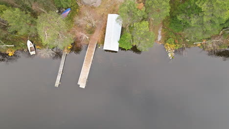Sweden---Autumn-Forest-Reflects-on-the-Water-by-a-Lakeside-on-a-Cloudy-Day,-Complemented-by-a-Floating-Wooden-Pier-and-Traditional-Fishermen's-Houses---Aerial-Sideways
