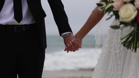 Bride-and-groom-hold-and-squeeze-tight-hands-with-sea-waves-in-background