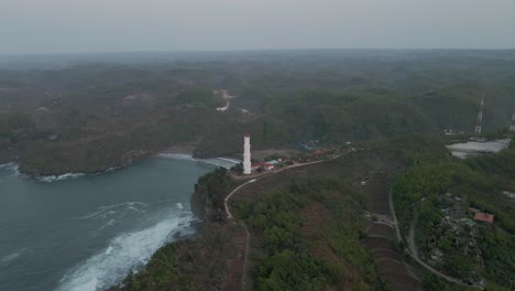 Aerial-view-of-white-lighthouse-on-the-hilly-shoreline-in-the-foggy-morning---Baron-beach,-Yogyakarta,-Indonesia