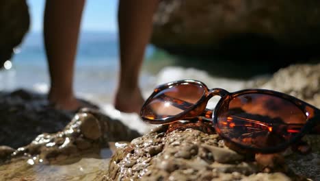 Sunglasses-on-beach-shore,-woman-going-swimming-in-the-sea-in-background