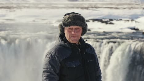 Man-look-into-camera,-massive-Godafoss-waterfall-in-background,-Iceland