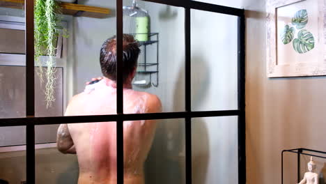 Caucasian-man-in-30s-washing-himself-with-soapy-sponge-in-home-shower,-back-view