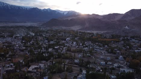 Vista-Aérea-Cinematográfica-De-La-Ciudad-De-Leh-Con-Vistas-A-La-Cordillera-Del-Himalaya-Cubierta-De-Nieve-Al-Fondo-Durante-La-Puesta-De-Sol,-Ladakh,-India.