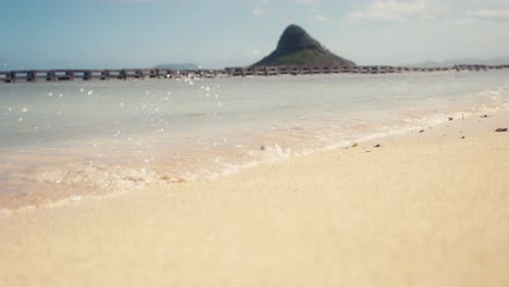 Nahaufnahme-Einer-Kleinen-Welle,-Die-An-Einem-Strand-Gegen-Den-Sand-Kracht,-Mit-Chinamans-Hat-Mokoliʻi-Im-Hintergrund-In-Oahu,-Hawaii