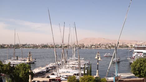 Schwenkaufnahme-Von-Booten,-Die-Entlang-Des-Nils-Treiben,-Mit-Bergen-Im-Hintergrund-Und-Segelbooten-Im-Vordergrund-In-Luxor,-Ägypten