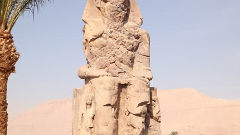 Ruinen-Der-Riesigen-Statue-Des-Pharaos-Mit-Bergen-Im-Hintergrund-Bei-Den-Kolosse-Von-Memnon-In-Luxor,-Ägypten-Mit-Palme
