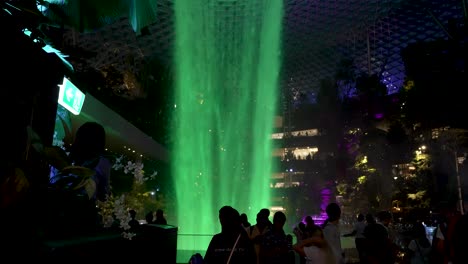 Hellgrün-Beleuchteter-Indoor-Wasserfall-Am-Jewel-Changi-Airport-Mit-Besuchern-Und-Passanten,-Die-Daneben-Fotos-Machen