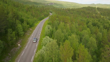 Campervan-drives-along-winding-mountain-road-lined-with-green-forest,-aerial