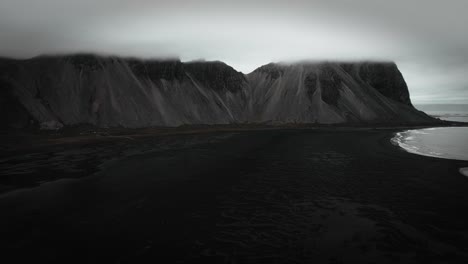 Aerial-black-sand-beach-stokksnes,-volcanic-dark-mountains,-vestrahorn-in-distance,-dark-moody-cloudy-scenery-Iceland