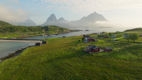 Holdoya-village-homes-on-grassy-slopes-above-ocean-with-jagged-mountains-wrapped-in-clouds,-Norway