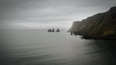 Cinematic-rock-formation-and-cliff-aerial-black-sand-beach-ocean-scenery,-moody-dark-scenery,-town-Vik,-Reynisdrangar-Iceland