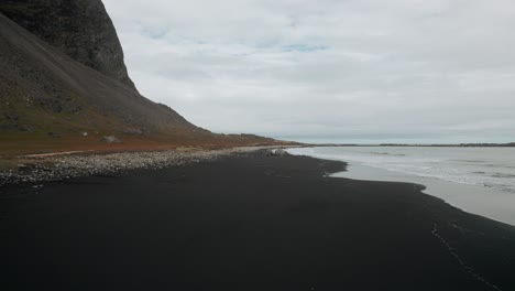Vuelo-Aéreo-A-Lo-Largo-De-La-Playa-De-Arena-Negra-Stokksnes,-Montañas-Volcánicas-Oscuras-En-La-Distancia,-Paisaje-Oscuro-Y-Nublado-Islandia