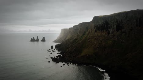 Cinematic-cliff-aerial-black-sand-beach-rock-formation,-ocean-scenery,-moody-dark-scenery,-town-Vik,-Reynisdrangar-Iceland