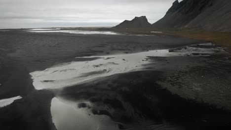 Playa-Aérea-De-Arena-Negra-Stokksnes,-Reflejos-De-Montañas-Volcánicas-Oscuras-En-La-Distancia,-Paisaje-Oscuro-Y-Nublado-Islandia