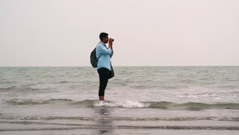 Male-photographer-standing-in-shallow-water-of-the-shore-facing-side-view-with-pants-curled-up-to-avoid-the-waves-taking-photos