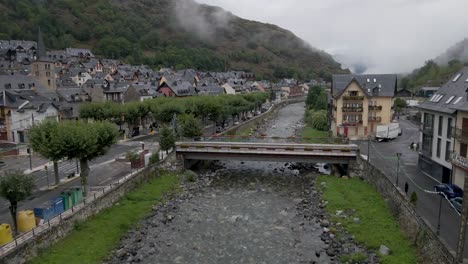 An-enchanting-drone-ascent-from-a-tranquil-riverbed-unveils-a-charming-Pyrenees-mountain-town-on-the-Spanish-side,-exuding-a-cozy-ambiance-on-a-cloudy,-cold-day