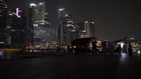 Silhouette-Von-Menschen,-Die-Nachts-An-Der-Promenade-Vorbeigehen,-Mit-Der-Skyline-Des-Finanzviertels-Von-Singapur-Im-Hintergrund