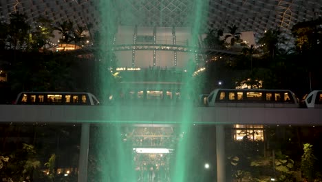 Der-Skytrain-Fährt-An-Einem-Indoor-Wasserfall-Vorbei,-Der-Von-Grünem-Licht-Am-Flughafen-Jewel-Changi-Beleuchtet-Wird