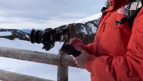 Man-with-red-snow-jacket-operates-professional-camera-mounted-on-stabilizing-gimbal-DJI-RS-3-Mini-surrounded-by-snowy-mountain-landscape