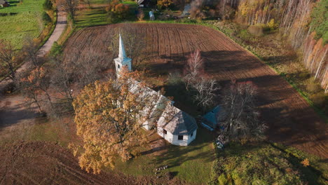 Rising-drone-shot-captures-historic-countryside-church-surrounded-by-fields,-forests-in-autumn-beauty