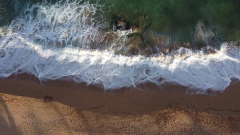Aerial-top-down-view-over-clear,-transparent-ocean-water-foaming-at-the-shore,-washing-the-golden-sand-of-the-beach