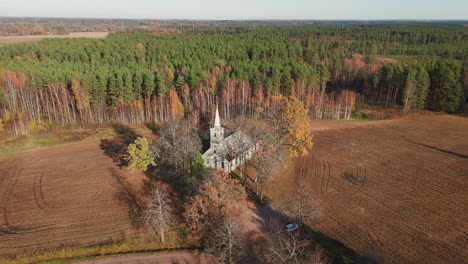 Drone-captures-Aerial-view-of-little-white-ancient-church-surrounded-by-empty-green-fields-and-forest-in-autumn-beauty
