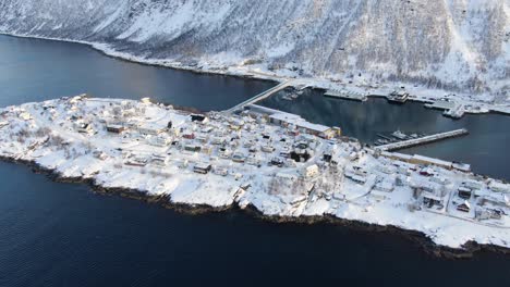 Drone-view-on-the-Tromso-mountains-in-winter-full-of-snow-showing-Husoy-a-small-town-on-an-island-surrounded-by-the-sea