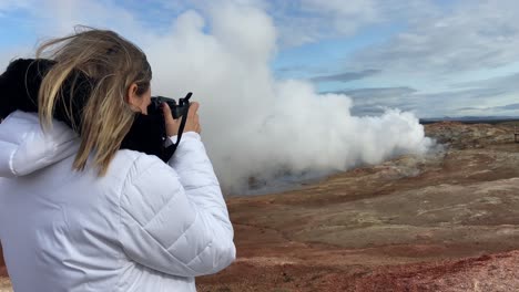 Mujer-Turista-Rubia-Tomando-Fotografías-Con-Un-Reflejo-De-Una-Fumarola-En-Gunnuhver,-Islandia