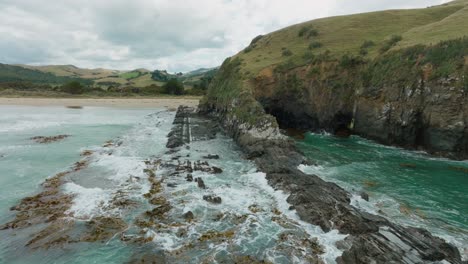 Aerial-drone-view-of-waves-breaking-on-rugged-and-rocky-coastline-covered-with-kelp-and-seaweed-on-the-remote-Cannibal-Bay,-Catlins,-South-Island-of-New-Zealand-Aotearoa