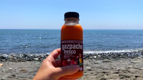 Holding-a-fresh-traditional-gazpacho-mini-bottle-from-Spanish-Mercadona-supermarket-at-the-sunny-beach-in-Estepona-Spain-with-sea-view,-cold-soup-on-the-go,-4K-shot