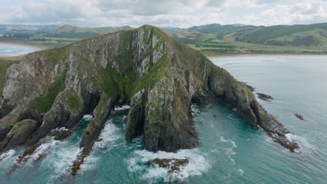 Aerial-view-of-rugged,-rocky-and-wild-landmass-and-crashing-waves-into-steep-cliffs-on-the-coastline-in-remote-wilderness-of-Cannibal-Bay,-Catlins,-South-Island-of-New-Zealand-Aotearoa