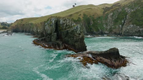 Aerial-drone-view-of-seaweed-kelp-covered-rocky-outcrop-and-steep-rugged-cliff-coastline-with-rough,-wild-ocean-of-Cannibal-Bay-in-the-Catlins,-South-Island-of-New-Zealand-Aotearoa