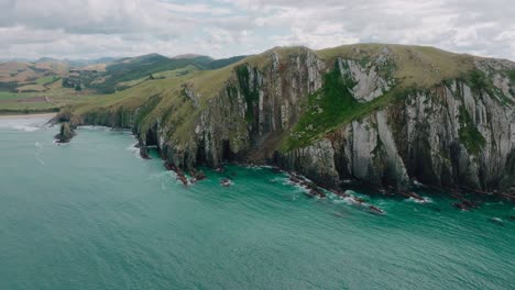 Aerial-landscape-view-of-steep,-rocky-and-rugged-cliff-coastline-with-turquoise-ocean-in-Cannibal-Bay-in-the-Catlins,-South-Island-of-New-Zealand-Aotearoa