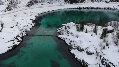Drone-view-in-Tromso-area-in-winter-flying-over-a-hanging-wooden-bridge-to-an-island-with-snow-with-a-clear-blue-sea-in-Norway