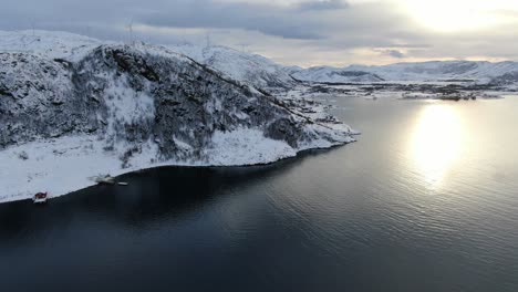Drohnenansicht-In-Der-Gegend-Von-Tromsø-Im-Winter,-Flug-über-Einen-Schneebedeckten-Berggipfel-Mit-Dem-Meer-Im-Vordergrund-Und-Windmühlen-Auf-Den-Bergen-In-Norwegen
