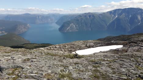 Spectacular-drone-shot-showing-mountain-biker-riding-on-rocky-summit-of-Norwegian-mountains---Beautiful-valley-with-blue-fjord-in-summer