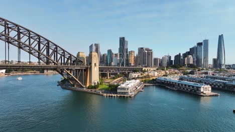 Aerial-Pullback-From-View-Of-Sydney-CBD-To-Reveal-Sydney-Harbour-Bridge-While-Ferry-Cruises-By-At-Sunset,-Australia