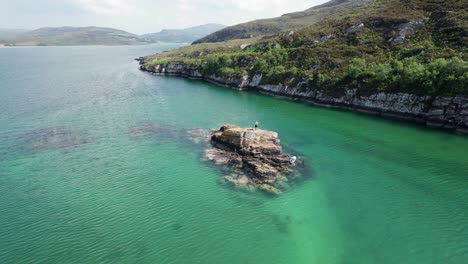 A-man-exploring-a-small-rocky-island-with-a-paddleboard-surrounded-by-turquoise-sea-water-in-a-loch-in-the-Scottish-highlands