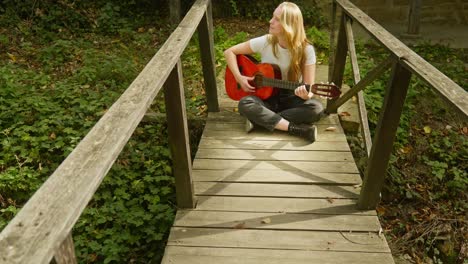 Junge-Schöne-Blonde-Frau-Spielt-Akustikgitarre-Auf-Einer-Holzbrücke