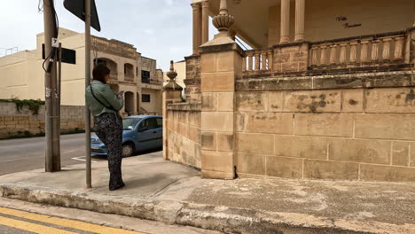 Mujer-Parada-En-La-Esquina-De-Una-Calle-En-Malta-Usando-Su-Teléfono-Y-Mirando-Hacia-La-Cámara