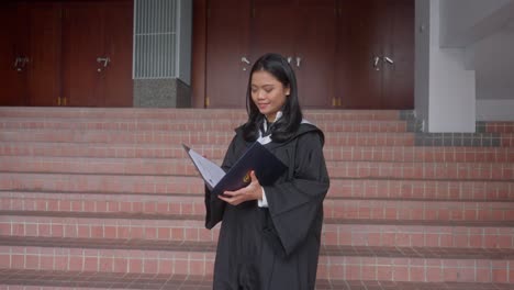 Chica-Asiática-Indonesia-Con-Toga-De-Graduación-Abriendo-Carpeta-Con-Certificado-Con-Orgullo
