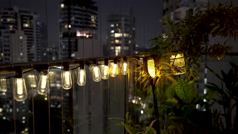 Row-Of-Dim-Lit-Bulbs-Hanging-Over-Balcony-Railing-With-Flickering-Light-Beside-Plants-At-Night-In-Singapore