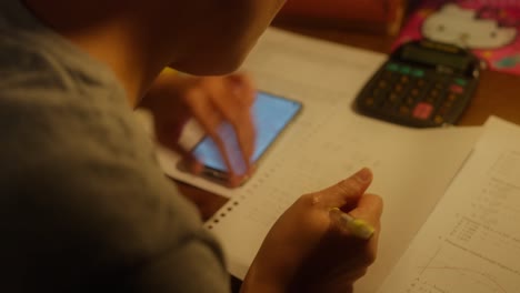 Closeup-of-Girl-Studying-Writing-Math-Homework-on-Desk-in-Room-With-Warm-Tungsten-Light-Using-Phone