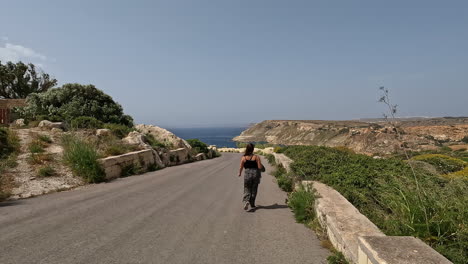 Mujer-Caminando-Por-Una-Carretera-En-Malta,-Europa-En-Un-Día-Ventoso-Y-Soleado-Con-El-Océano-Al-Fondo