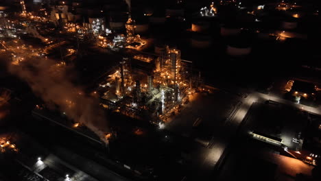 Large-oil-refinery-plant-with-thick-smoke-coming-from-chimneys,-aerial-night-establishing-view