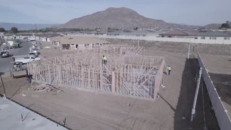 A-drone-progressively-approaches-a-partially-built-house-with-only-raised-wall-frames,-highlighting-a-construction-worker-in-the-center-diligently-nailing-all-the-frames-together
