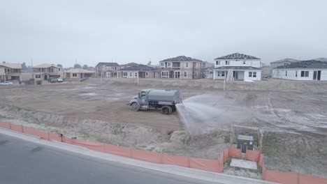 A-drone-observes-a-water-truck-as-it-diligently-dumps-water-on-a-dirt-lot-within-a-home-construction-site,-facilitating-the-site's-preparation
