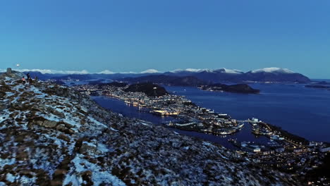 Aerial-view-over-a-snowy-mountain,-revealing-the-Alesund-city,-blue-hour-in-Norway